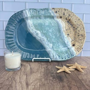 May include: An oval ceramic platter featuring a gradient of deep blue, light turquoise, and sandy beige with a textured, rippled edge. A white candle in a glass jar and two small, light-colored starfish are positioned in front of the platter.
