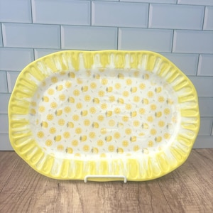 May include: A yellow ceramic serving platter with a scalloped edge. The interior features a pattern of yellow lemons on a white background. The platter is oval-shaped and rests on a white metal stand.