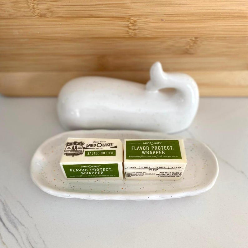 May include: A white ceramic whale-shaped butter dish with a speckled oval tray holding two Land O Lakes butter packets. One packet reads "Salted Butter" and the other reads "Flavor Protect Wrapper".
