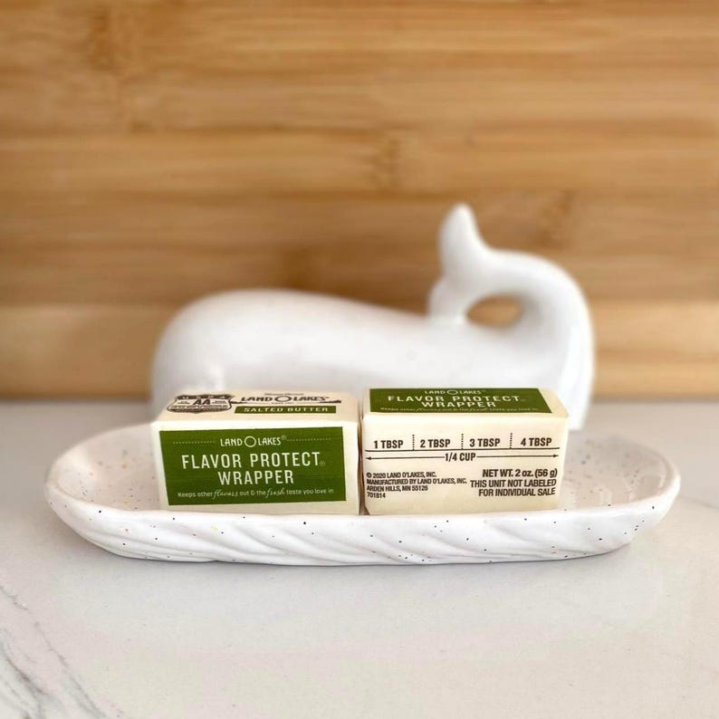 May include: A white ceramic whale-shaped butter dish holds two wrapped butter portions. The butter wrappers are green and white, with the text "Flavor Protect Wrapper" and measurement markings. The dish sits on a white surface.