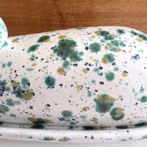 May include: White ceramic butter dish with a speckled design in shades of green, gold, and blue. The dish has a rounded top and sits on a matching base. The background is a wooden surface.