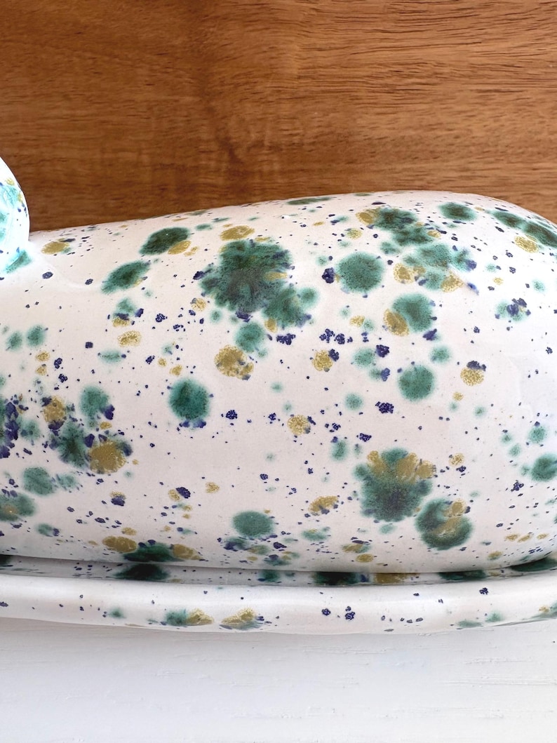 May include: White ceramic butter dish with a speckled design in shades of green, gold, and blue. The dish has a rounded top and sits on a matching base. The background is a wooden surface.
