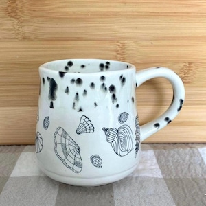 May include: White ceramic mug with a black speckled rim and handle. The mug features black line drawings of seashells. The mug is on a checkered surface and a wooden background.