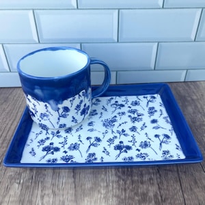 May include: A ceramic mug and a matching rectangular tray featuring a white base with a blue floral pattern. The mug has a solid blue upper section with a drip-like glaze effect, while the tray has a solid blue border framing the floral design.