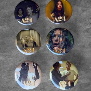 May include: Six circular pin-back buttons featuring various film scenes and the A24 logo in gold lettering. The designs include portraits of individuals, figures in a hallway, and a person with a camera, all rendered in a glossy, high-resolution finish.