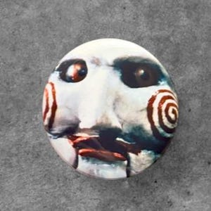 May include: A circular pinback button featuring a graphic illustration of a pale, white-faced puppet with dark eye sockets, a prominent nose, and distinct red spiral patterns on each cheek, set against a stark, high-contrast background.
