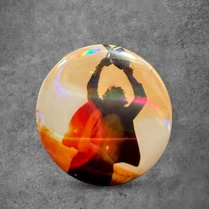 May include: A circular pinback button featuring a silhouette of a person with arms raised above their head. The background of the design displays a warm gradient of orange, yellow, and white tones with a subtle iridescent sheen across the surface.