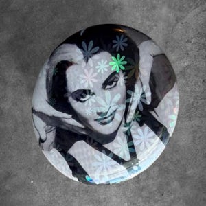 May include: A circular glass paperweight featuring a black and white photographic portrait of a person with dark hair, overlaid with a repeating pattern of small, light-colored, and iridescent flower shapes that catch the light.