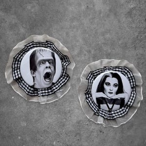 May include: Two circular pin-back buttons featuring black and white portraits of a male monster and a female character. Each button is framed by a ruffled beige fabric border and a black and white checkered ribbon trim, creating a layered effect.