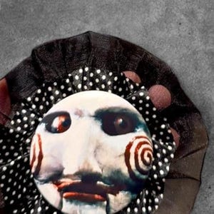 May include: A circular pinback button featuring a pale, white-faced character with dark eye sockets and red spiral cheek markings. The button is framed by a ruffled, black fabric border with a white polka dot pattern and a sheer black outer layer.