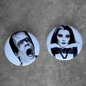 May include: Two circular pin-back buttons featuring black and white portraits. The left button displays a close-up of a man with a flat-top hairstyle and a wide-open mouth. The right button shows a woman with long dark hair and light streaks.