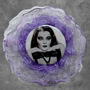 May include: A circular pinback button featuring a black and white portrait of a person with long dark hair, resting their chin on their hands. The button is surrounded by layers of ruffled, sheer light purple and white lace fabric trim.