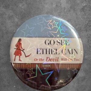 May include: A round pinback button featuring a holographic star overlay. The design shows a person in a red dress holding a scythe, with the text: GO SEE ETHEL CAIN Or the Devil Will Get You! and a small Lamar logo on a brown horizontal band.