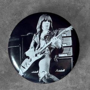 May include: A circular pin-back button featuring a black and white photograph of a musician with long hair wearing a leather jacket and playing an electric guitar. The background shows stacked Marshall amplifiers with the brand name clearly visible.