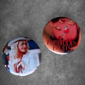 May include: Two circular pin-back buttons. One button features a person with blonde hair wearing a white hat and a white patterned top. The other button is red with gold-toned fruit charms and the text FORBIDDEN FRUITS printed in a bold red font.