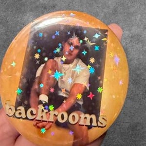 May include: A round pinback button featuring a holographic star overlay on a photograph of a person wearing a white t-shirt. The word backrooms is printed in a bold, sans-serif font across the bottom of the circular button in a light tan color.