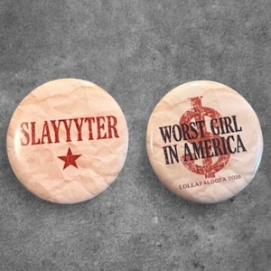 May include: Two round pinback buttons with a light beige, wrinkled paper texture. The left button features the red text SLAYYYTER above a red star. The right button displays the black text WORST GIRL IN AMERICA over a red dollar sign symbol.