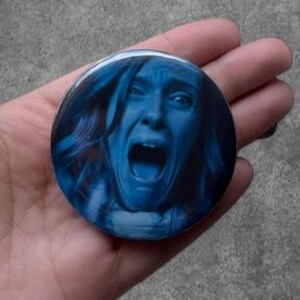 May include: A circular pin-back button featuring a monochromatic blue-toned photographic print of a person with long hair and an open mouth in a screaming expression. The image is centered on the round button, which has a glossy finish.
