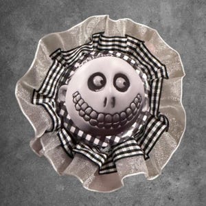 May include: A circular decorative accessory featuring a central white character face with large black eyes and a wide toothy grin. The face is framed by a ruffled border of black and white checkered fabric, surrounded by an outer layer of shimmering silver fabric.