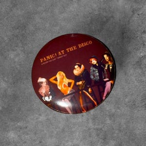 May include: A circular pinback button featuring the Panic! At The Disco album cover for A Fever You Can't Sweat Out. The dark red background displays the band name and album title in gold text above four figures in vintage-style clothing.