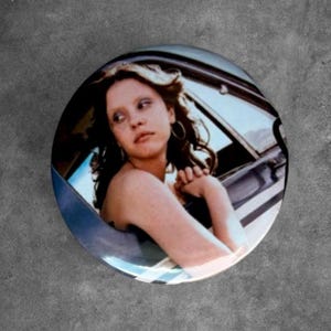 May include: A circular pinback button featuring a photographic print of a person with long wavy hair and hoop earrings, looking over their shoulder while sitting inside a vehicle. The image captures a candid moment with soft lighting.