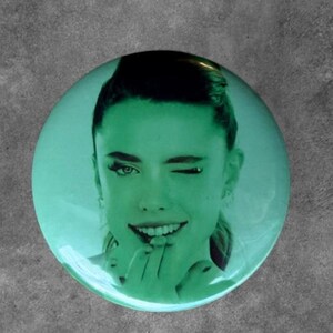 May include: A circular pinback button featuring a monochromatic green-tinted photographic portrait of a person with their hair pulled back. The person is winking with one eye and has a finger placed near their mouth in a playful, expressive gesture.