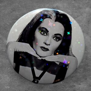 May include: A circular pinback button featuring a black and white portrait of a person with long dark hair and a white streak, resting their chin on their hands. The surface has a holographic finish with small colorful star-shaped light reflections.