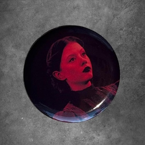 May include: A circular decorative plate featuring a high-contrast portrait of a person with dark hair and dark lipstick. The image is bathed in deep red lighting against a black background, highlighting the facial features and shoulders.