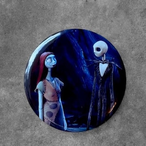 May include: A circular pin featuring a graphic illustration of two characters against a dark blue background. On the left, a female character with long red hair and a tan dress. On the right, a tall, thin, white skeletal character in a black suit.
