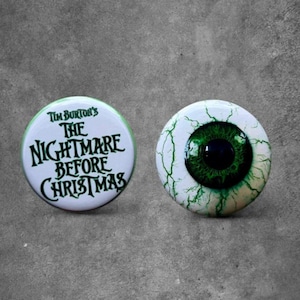 May include: Two circular pin-back buttons. One button features the white text Tim Burton's The Nightmare Before Christmas in a stylized black font on a white background. The other button displays a realistic human eyeball with a green iris.