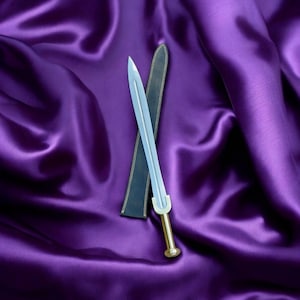 May include: A small, silver sword with a gold handle and a black sheath lies on deep purple satin. The sword has a straight, double-edged blade and a rounded pommel. The sheath is open, revealing the sword.