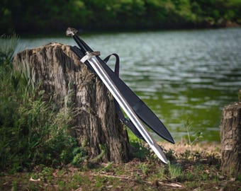 May include: A silver and black sword with a decorative handle rests against a tree stump. The sword is sheathed in a black scabbard with a carrying strap. The background features a lake and greenery.