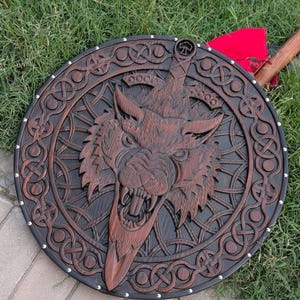 Könnte beinhalten: Ein runder Holzschild mit einem tief geschnitzten Wolfskopf in der Mitte, der in ein Schwertdesign integriert ist. Der Schild ist mit komplizierten keltischen Knotenmustern am Rand verziert und mit silberfarbenen Metallnieten versehen.