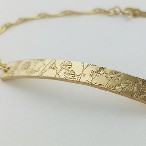 May include: A gold bracelet with a delicate chain and a curved bar charm featuring a floral design.