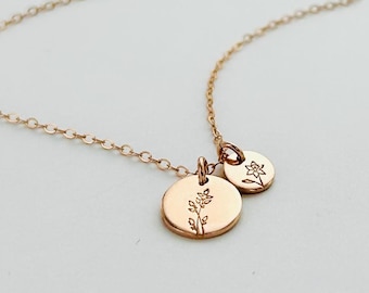 May include: A delicate gold-colored necklace featuring two small, round pendants. One pendant displays a floral design, while the other has a smaller floral motif. The necklace chain is fine and elegant, perfect for everyday wear.