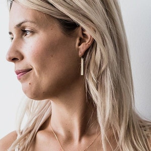 May include: A woman wearing gold bar earrings. The earrings are simple and elegant, with a minimalist design.