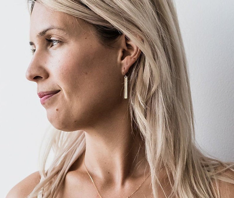 May include: A woman wearing gold bar earrings. The earrings are simple and elegant, with a minimalist design.