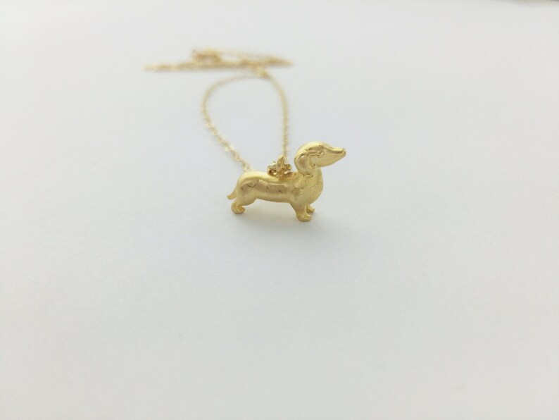 May include: A gold necklace with a dachshund pendant. The dachshund is standing on all four legs and has a long snout.
