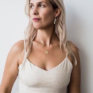 May include: A woman with blonde hair wearing a light beige linen dress with a gold necklace and earrings. The necklace has a round pendant with a textured surface.