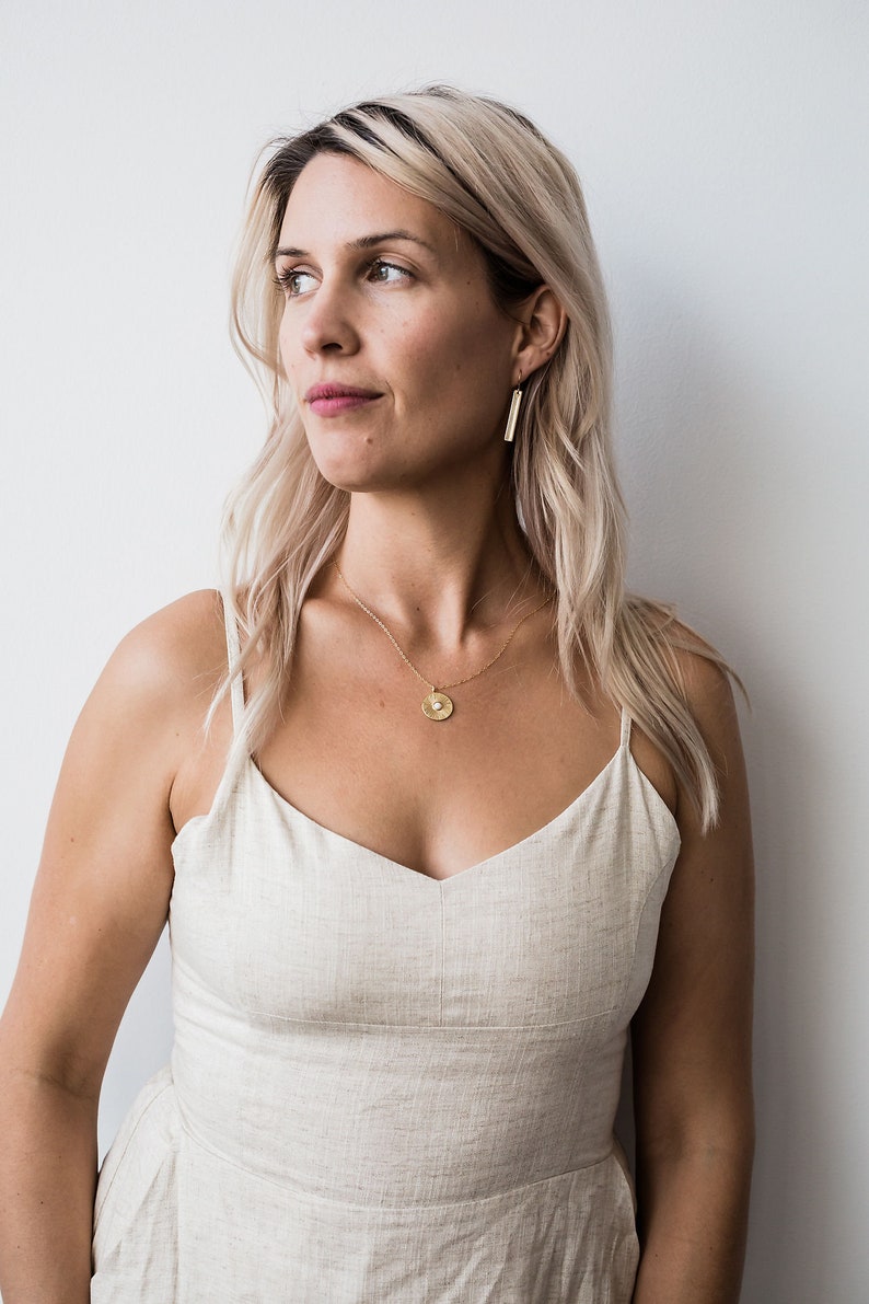 May include: A woman with blonde hair wearing a light beige linen dress with a gold necklace and earrings. The necklace has a round pendant with a textured surface.