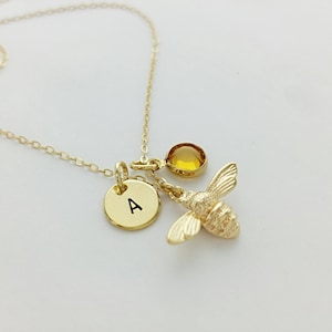 May include: Gold necklace with a bee charm, a small round charm with the letter A, and a yellow gemstone.