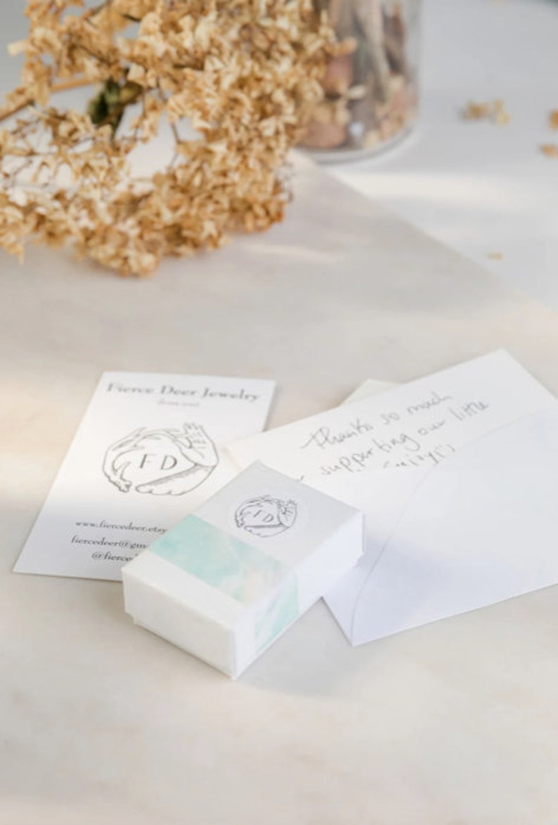 May include: A white box with a watercolor design and the letters "FD" in a circle. The box is sitting on a white surface next to a white card with the text "Fierce Deer Jewelry" and "Thanks so much supporting our little mimi!"