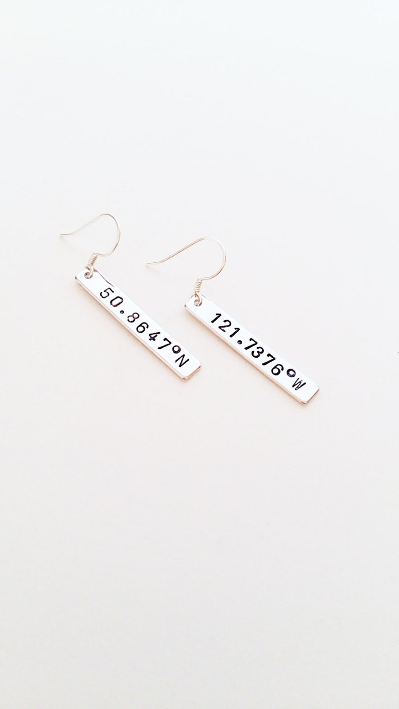May include: A pair of silver earrings with rectangular charms. The charms have the coordinates "50.8647&deg;N" and "121.7376&deg;W" engraved on them.