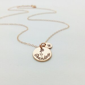 May include: Rose gold necklace with a pendant that says "Grandma" and a small flower illustration. A small charm with the letter "A" is also attached. The necklace is on a white background.