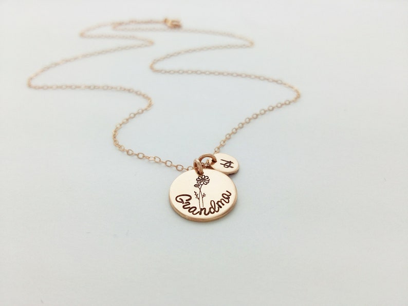May include: Rose gold necklace with a pendant that says "Grandma" and a small flower illustration. A small charm with the letter "A" is also attached. The necklace is on a white background.