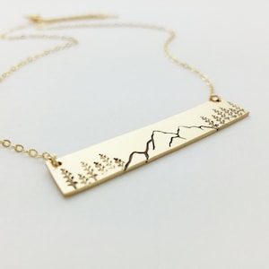May include: A gold necklace with a rectangular pendant featuring a minimalist line drawing of mountains and trees.