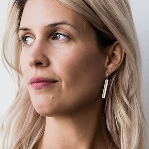 May include: A woman with blonde hair wearing a gold bar pendant necklace and gold bar earrings.