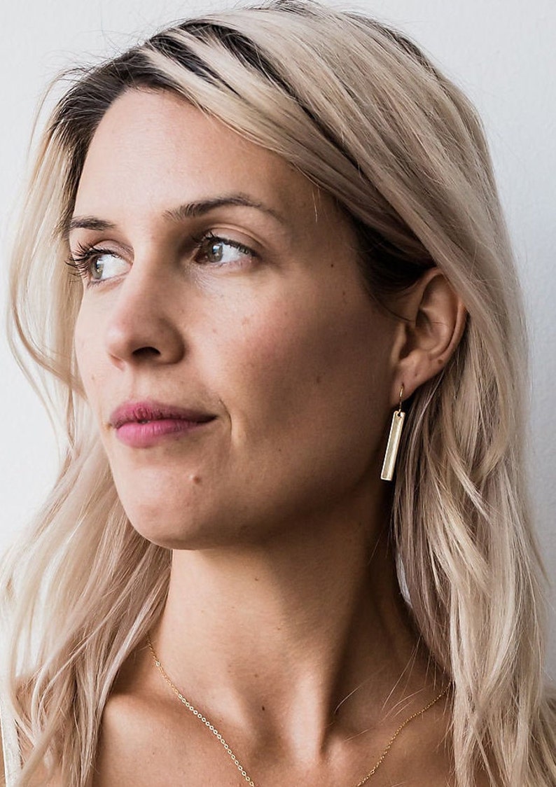 May include: A woman with blonde hair wearing a gold bar pendant necklace and gold bar earrings.