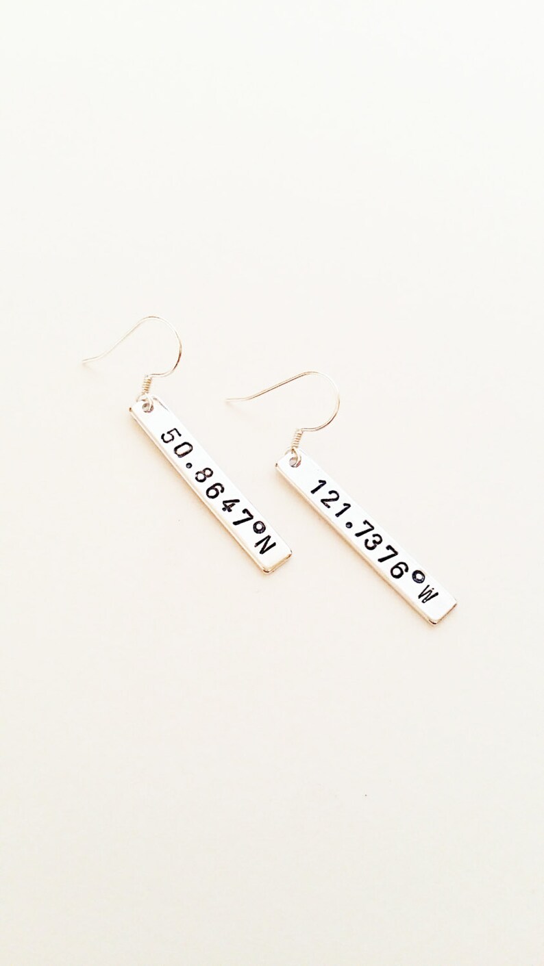May include: A pair of silver earrings with rectangular charms. The charms have the coordinates "50.86470N" and "121.73760W" engraved on them.