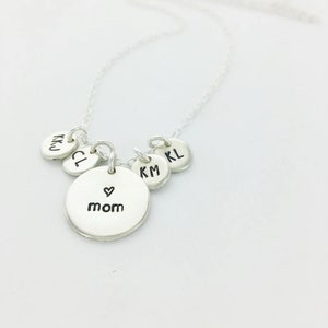 May include: A silver necklace with a delicate chain. The necklace features a central round pendant with the word "mom" and a heart symbol, along with smaller round pendants with initials. The necklace is displayed against a white background.